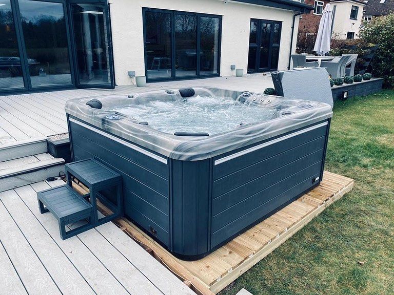 Hot tub on wooden deck, next to a house with large windows. Grey steps lead up to the tub.