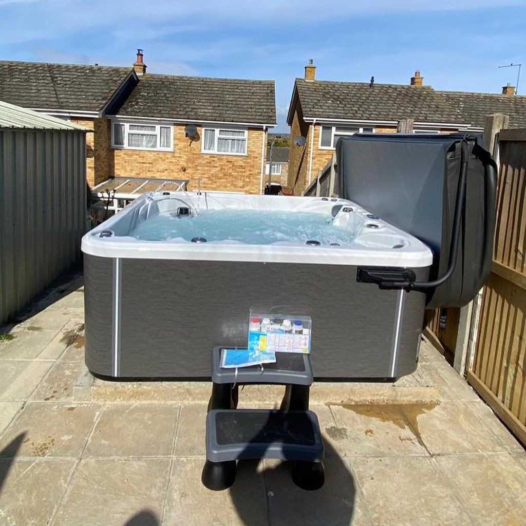 Hot tub in a backyard setting, partially open cover, gray and blue tones, step stool in front.