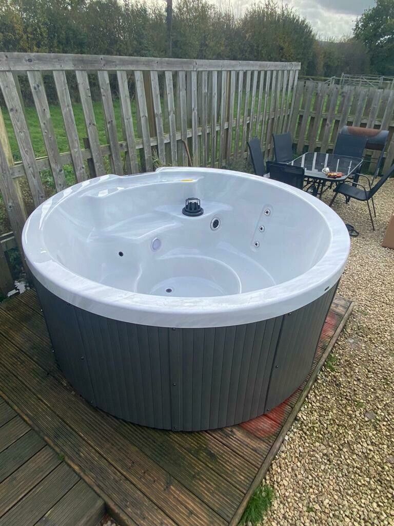 Round hot tub with gray exterior on a wooden deck in front of a fence and patio furniture.