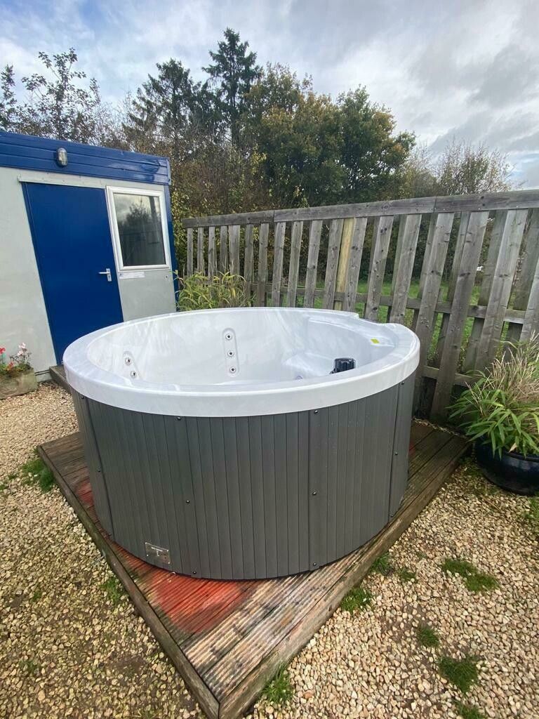 Round hot tub with gray siding sits on wooden platform near fence and small shed.