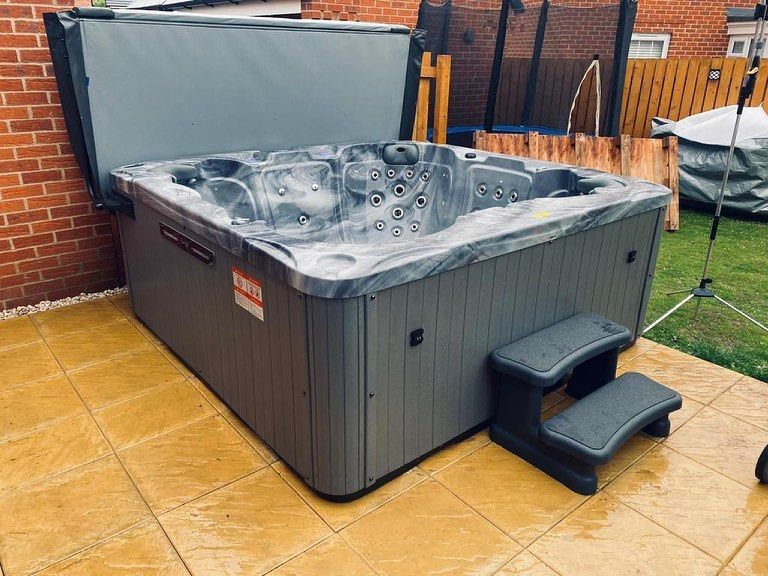 Gray hot tub with the cover open, steps, on a patio, next to a brick wall.