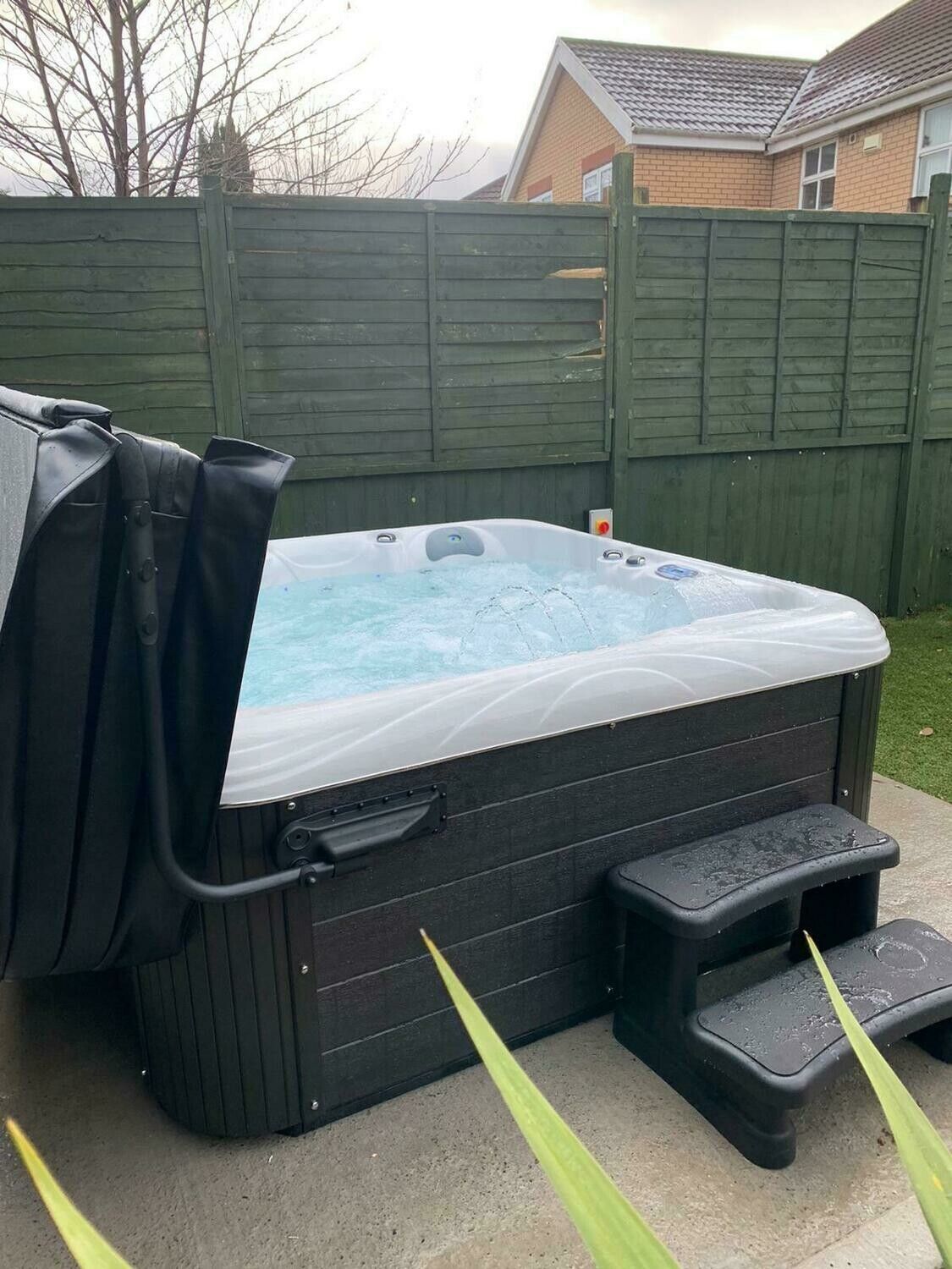 Hot tub with cover partially open, black steps beside it. Outdoor setting with green fence and overcast sky.