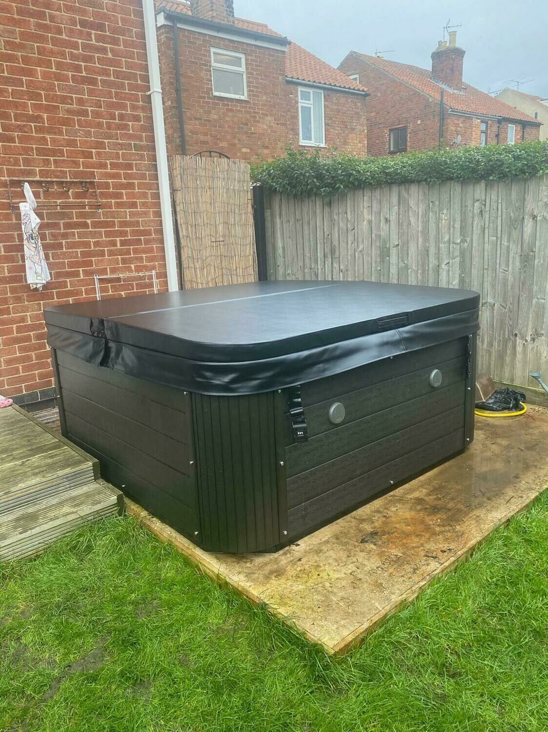 Black hot tub with closed cover on concrete in a backyard, beside brick and wooden fences.
