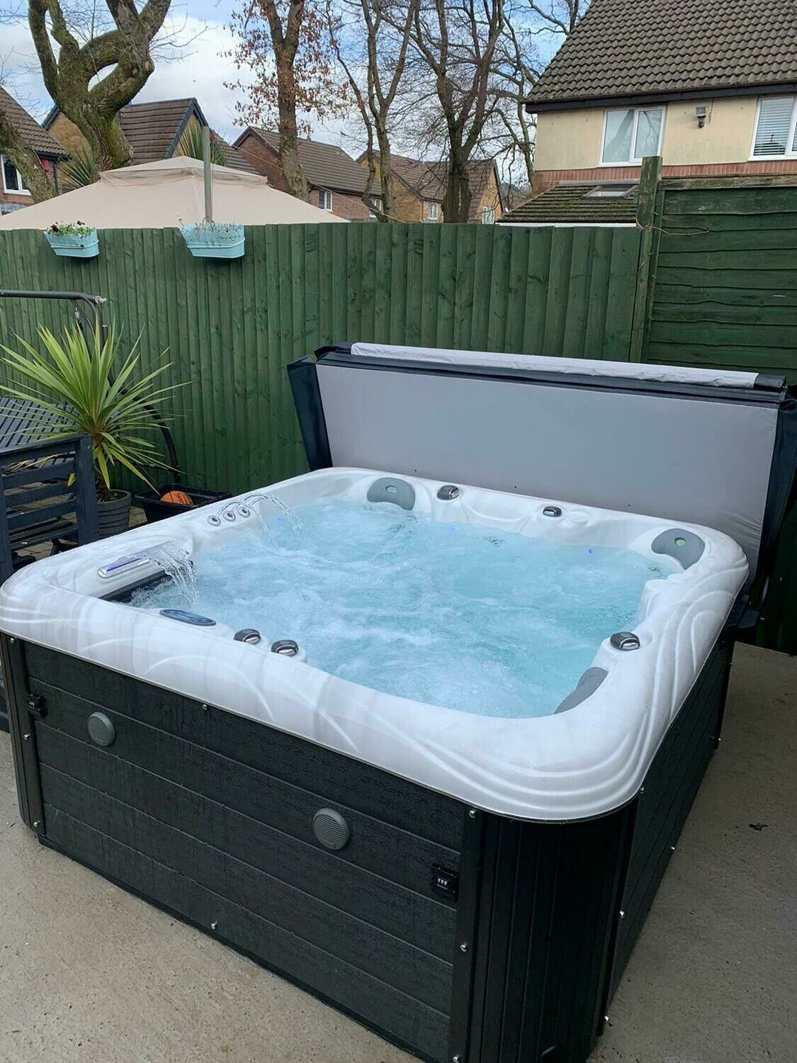 Hot tub with open cover, filled with water, set in a backyard with a green fence and beige umbrella.