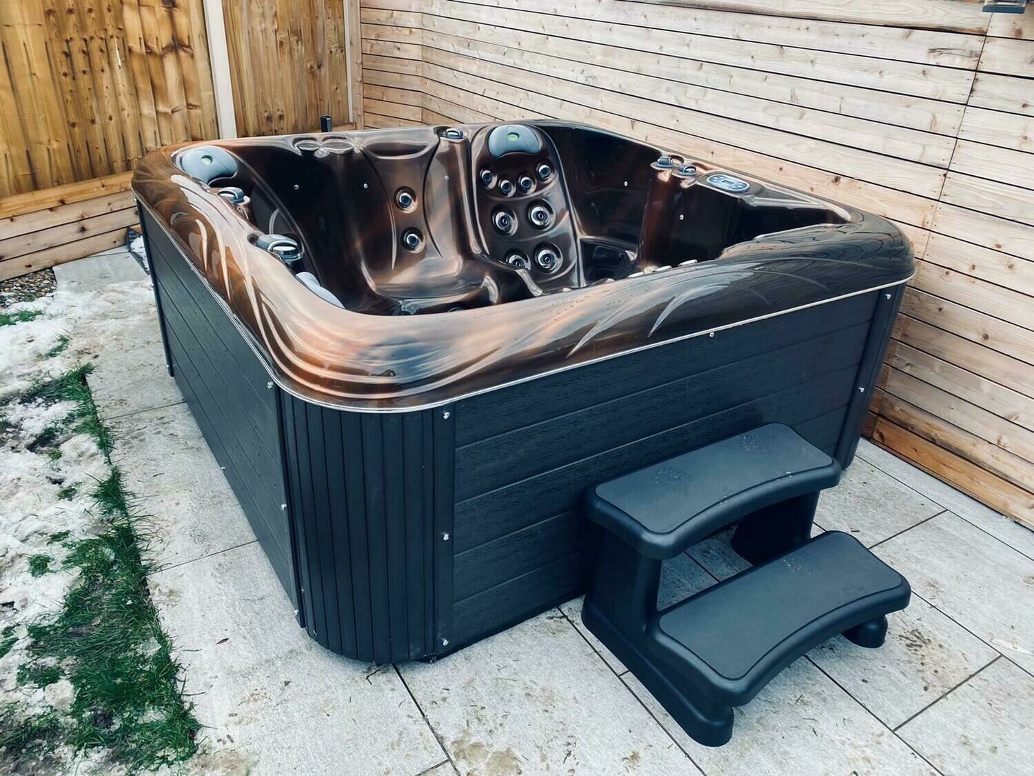 Hot tub with dark brown interior, black exterior, and step stool on a tiled patio.