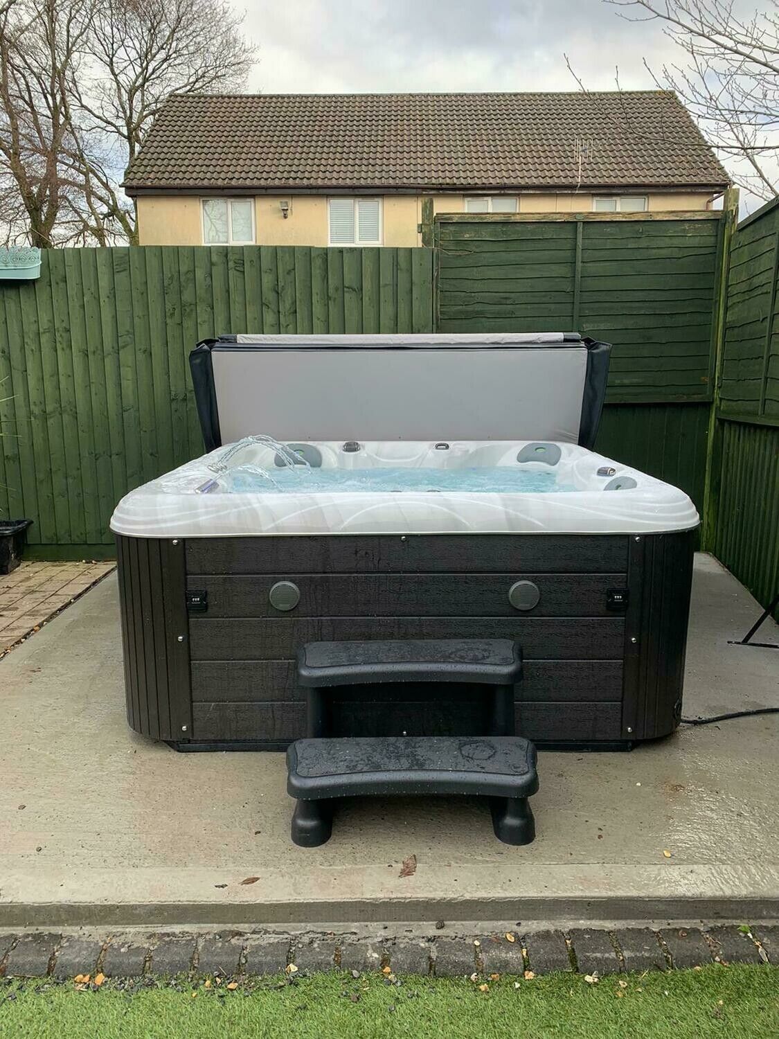 Hot tub with open cover, dark gray siding, black steps, in a backyard.