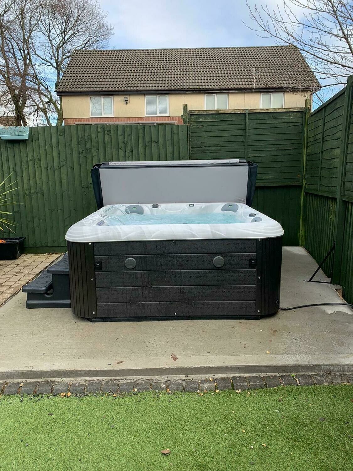 Hot tub with open cover, on a concrete patio, next to a green fence.