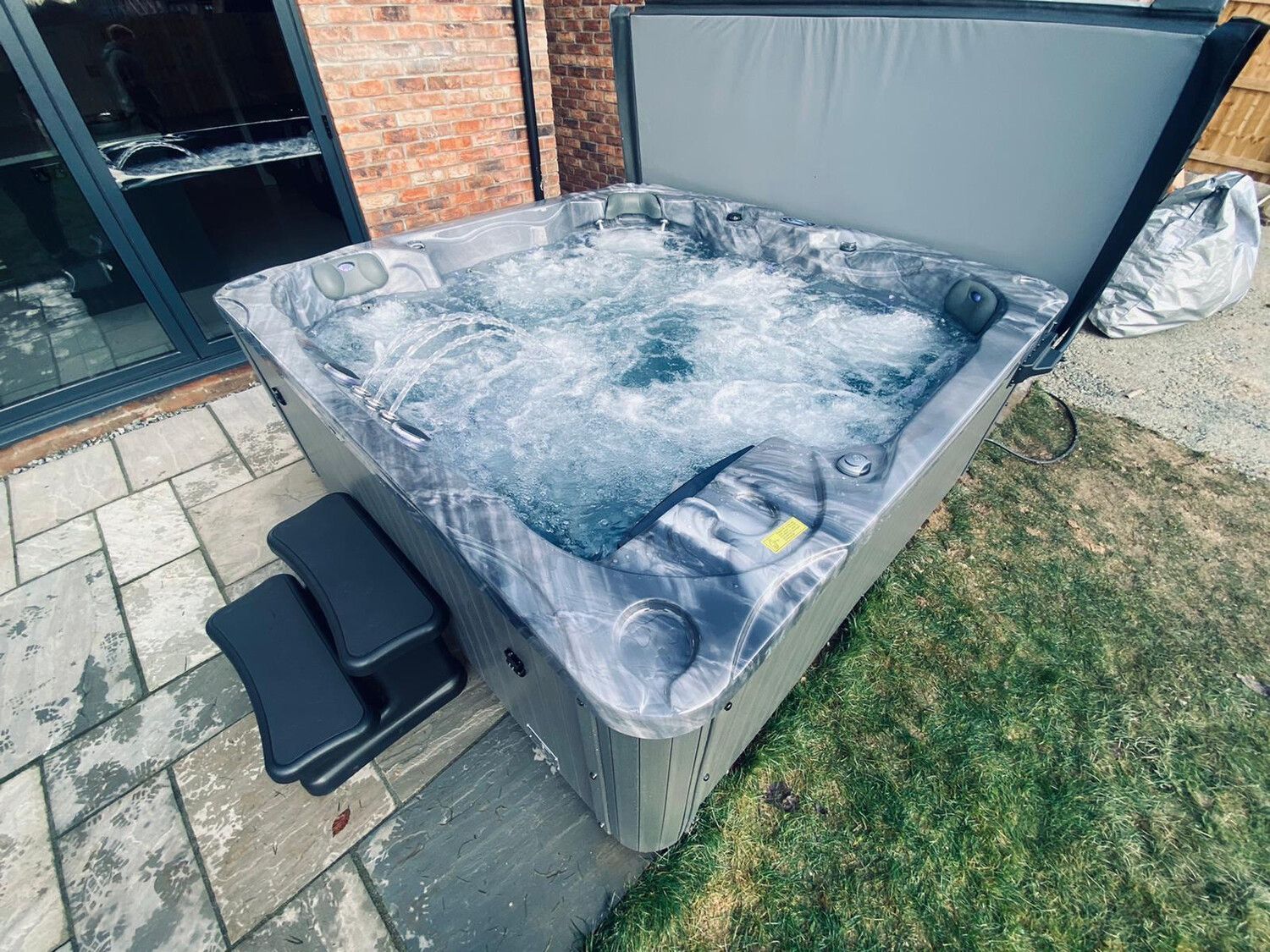 Hot tub bubbling on a patio, steps to access. Cover partially open. Green grass and brick building in background.