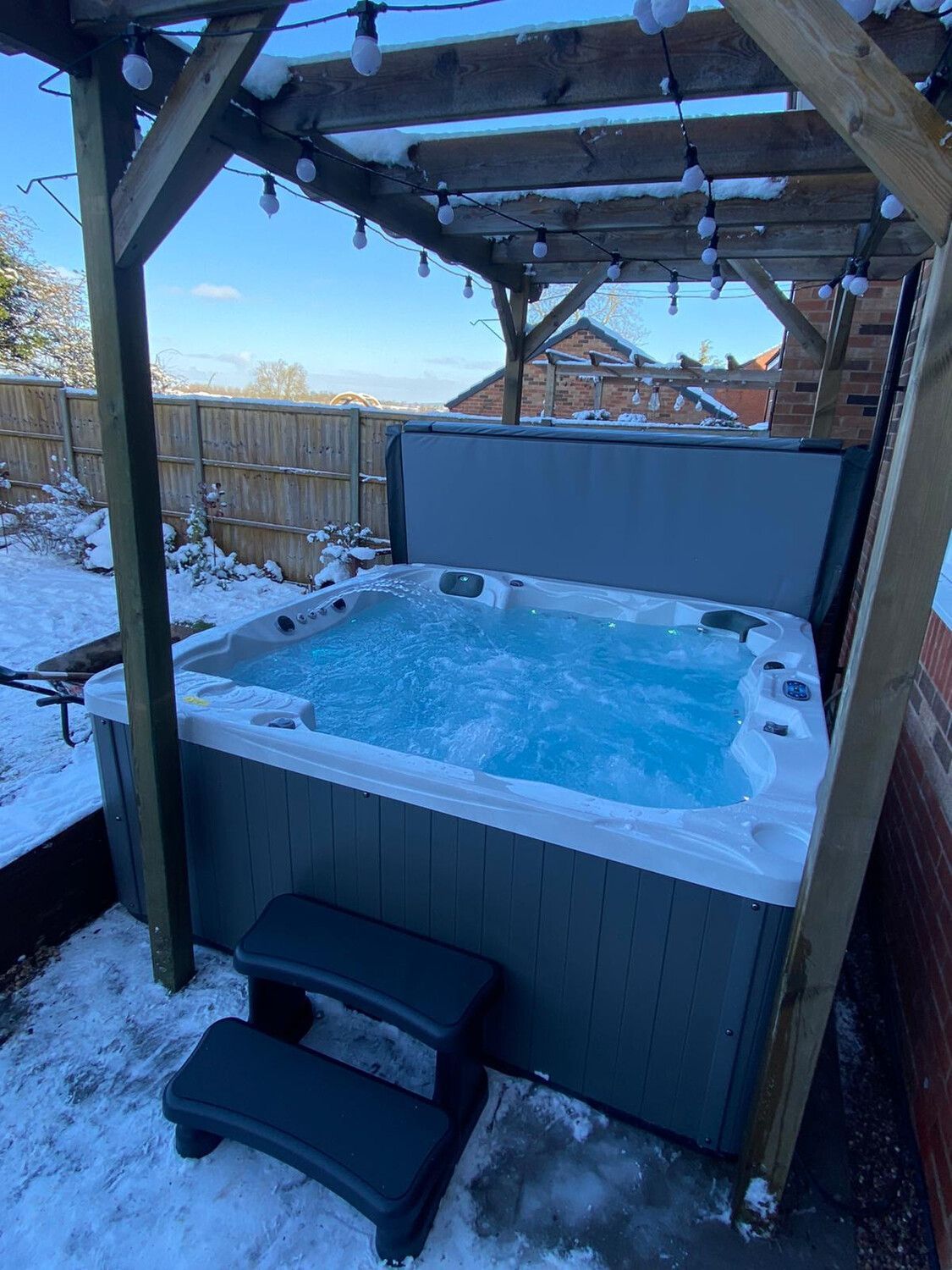 Hot tub with bubbling blue water, in a snowy outdoor setting under a wooden pergola.