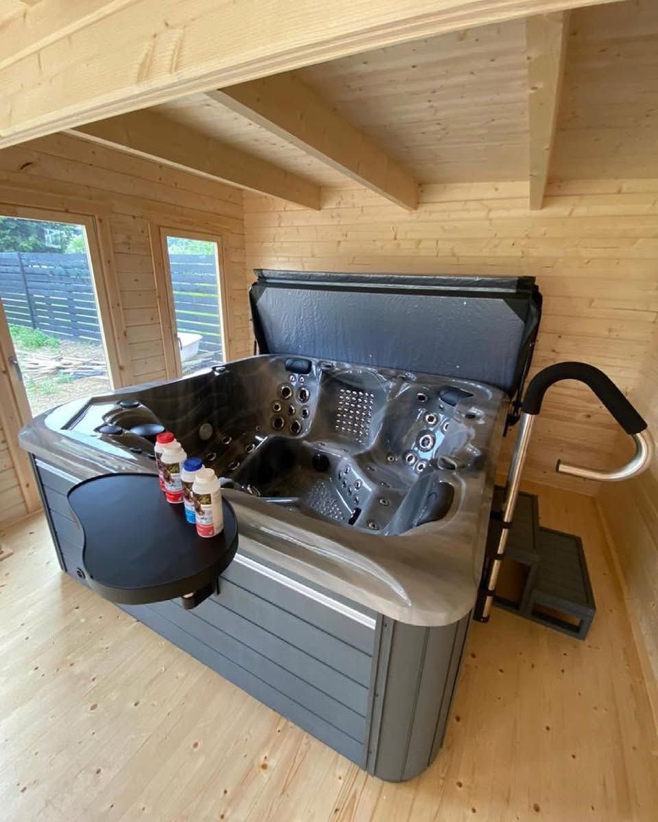 Hot tub in a wooden cabin.  Black tub cover open, chemical bottles on the side, stairs with a handrail.