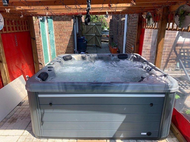 A hot tub sits under a wooden pergola. Bubbles rise in the water. Brick walls and a doorway are in the background.