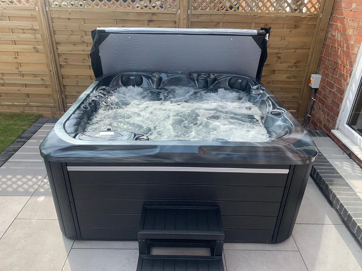 Black hot tub with grey cover open, bubbling water, set on gray patio, against a wooden fence.