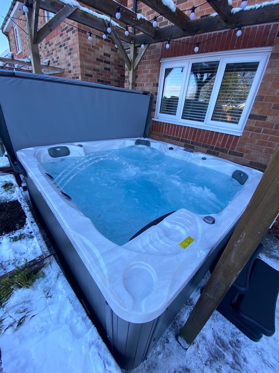 A rectangular hot tub with bubbling water sits outside in the snow, next to a brick wall and a window.