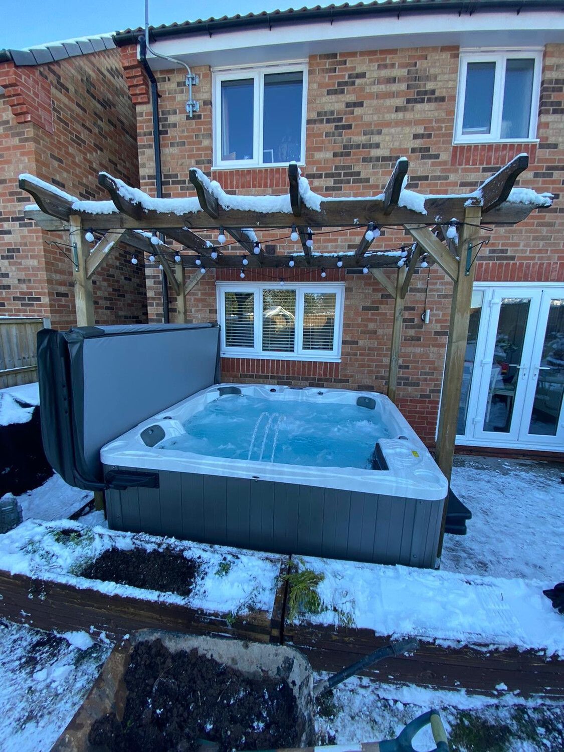 Hot tub with partially open cover under a snow-covered pergola, surrounded by snow in a backyard setting.