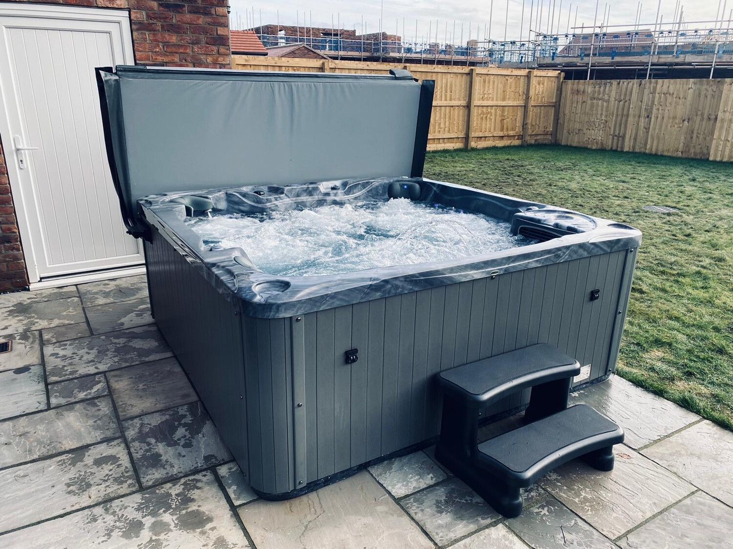 Hot tub with open cover, bubbling water, gray siding, black steps on a patio.