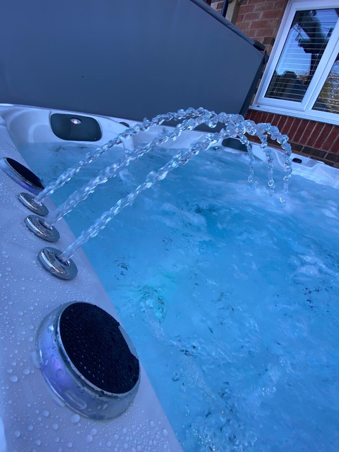 Hot tub with water jets spraying into clear blue water. Close up with gray cover and home exterior.