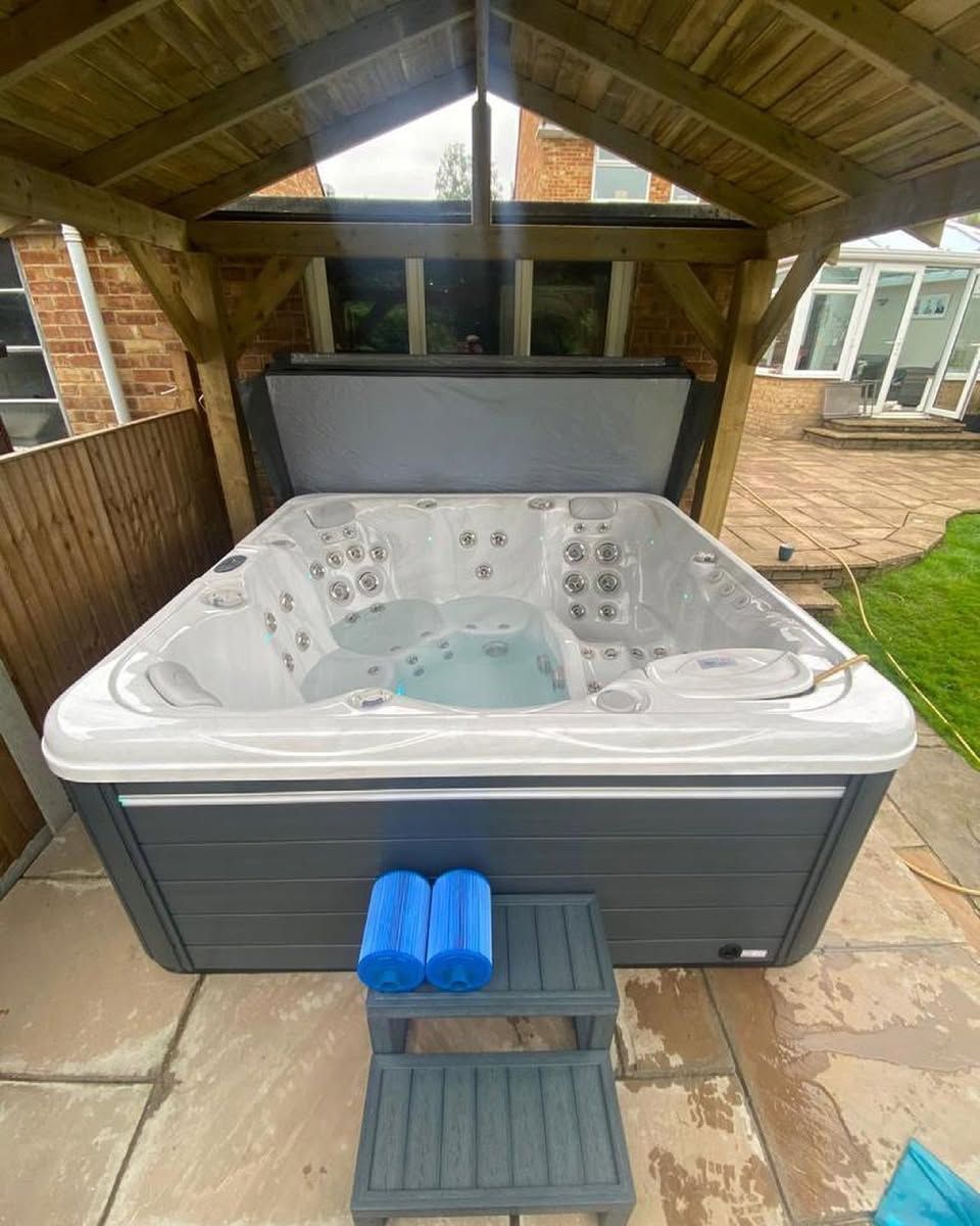 Hot tub under wooden pergola on a stone patio, with blue pool noodles and a step.