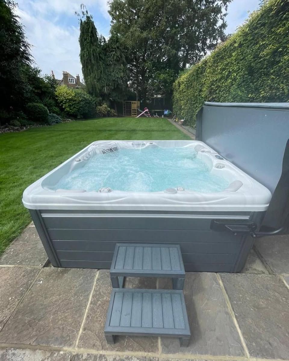 Hot tub on a stone patio with steps, surrounded by a green lawn and hedge.