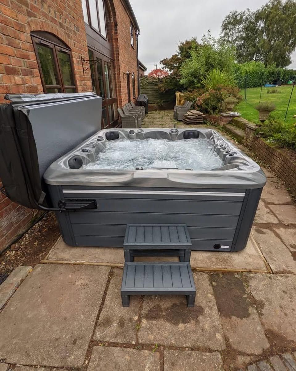 A dark gray hot tub with bubbling water sits on a stone patio, beside brick house, with a step.