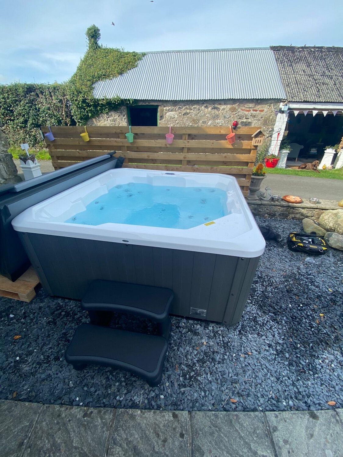 Hot tub with steps on a stone patio, with a wooden fence and a stone building in the background.