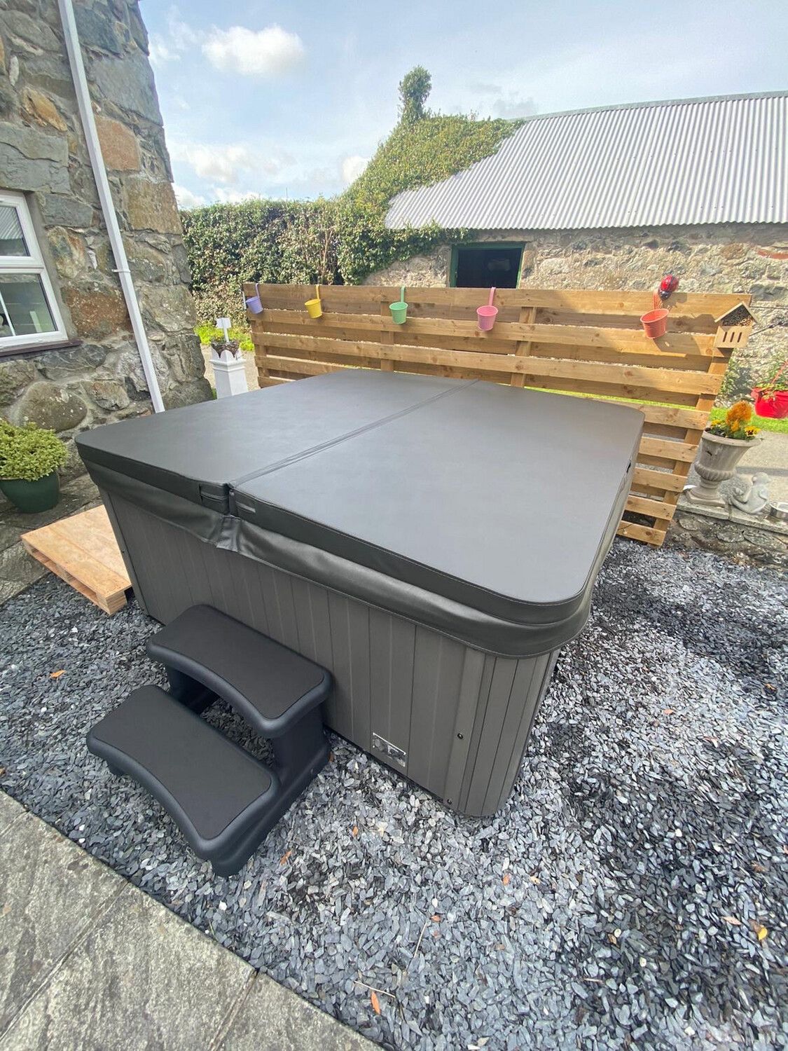 Hot tub with closed cover and steps in a stone courtyard. Wooden fence and ivy-covered building in the background.