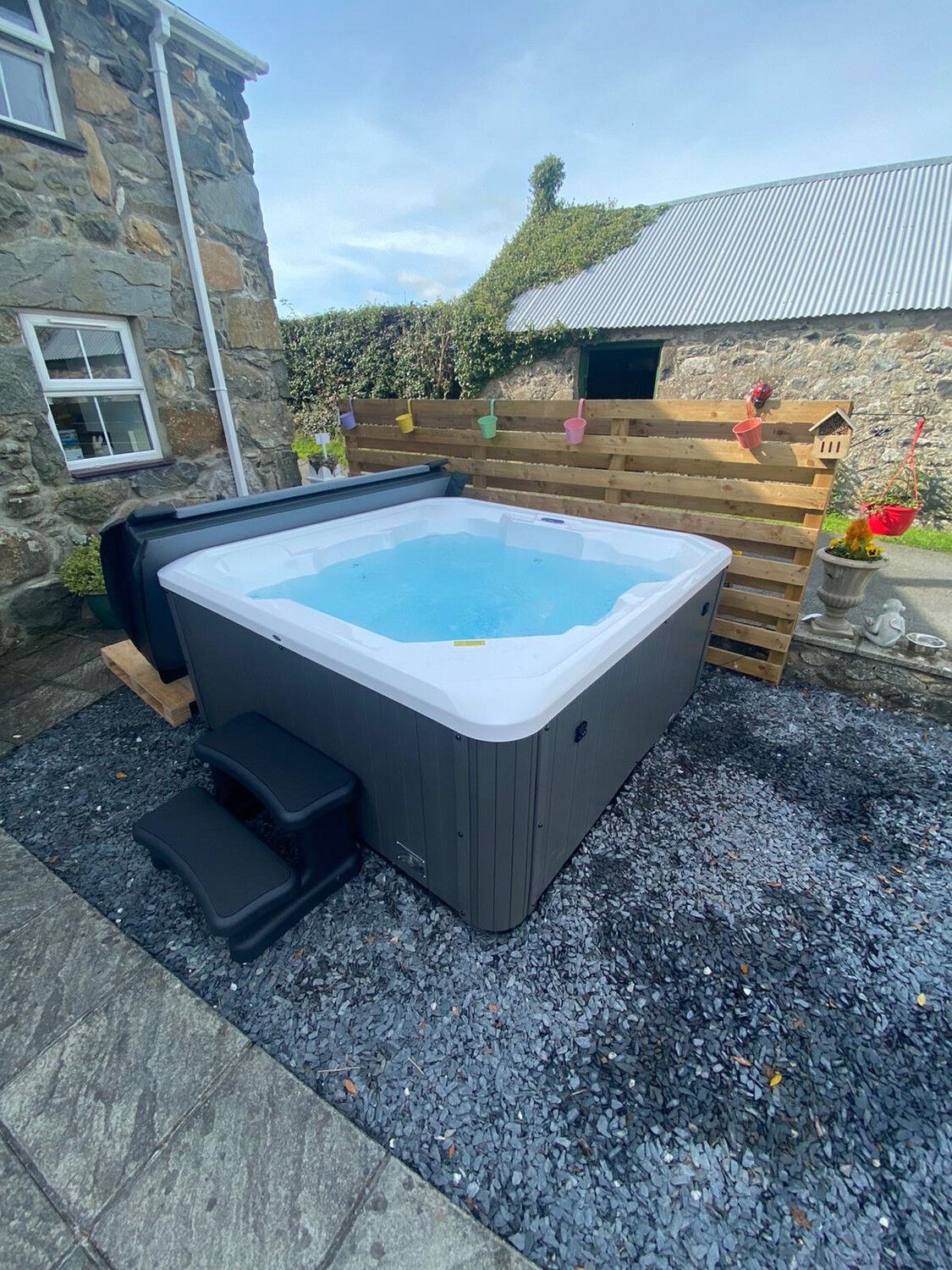 Hot tub on a gravel patio beside a stone building, with wooden fencing and a barn in the background.