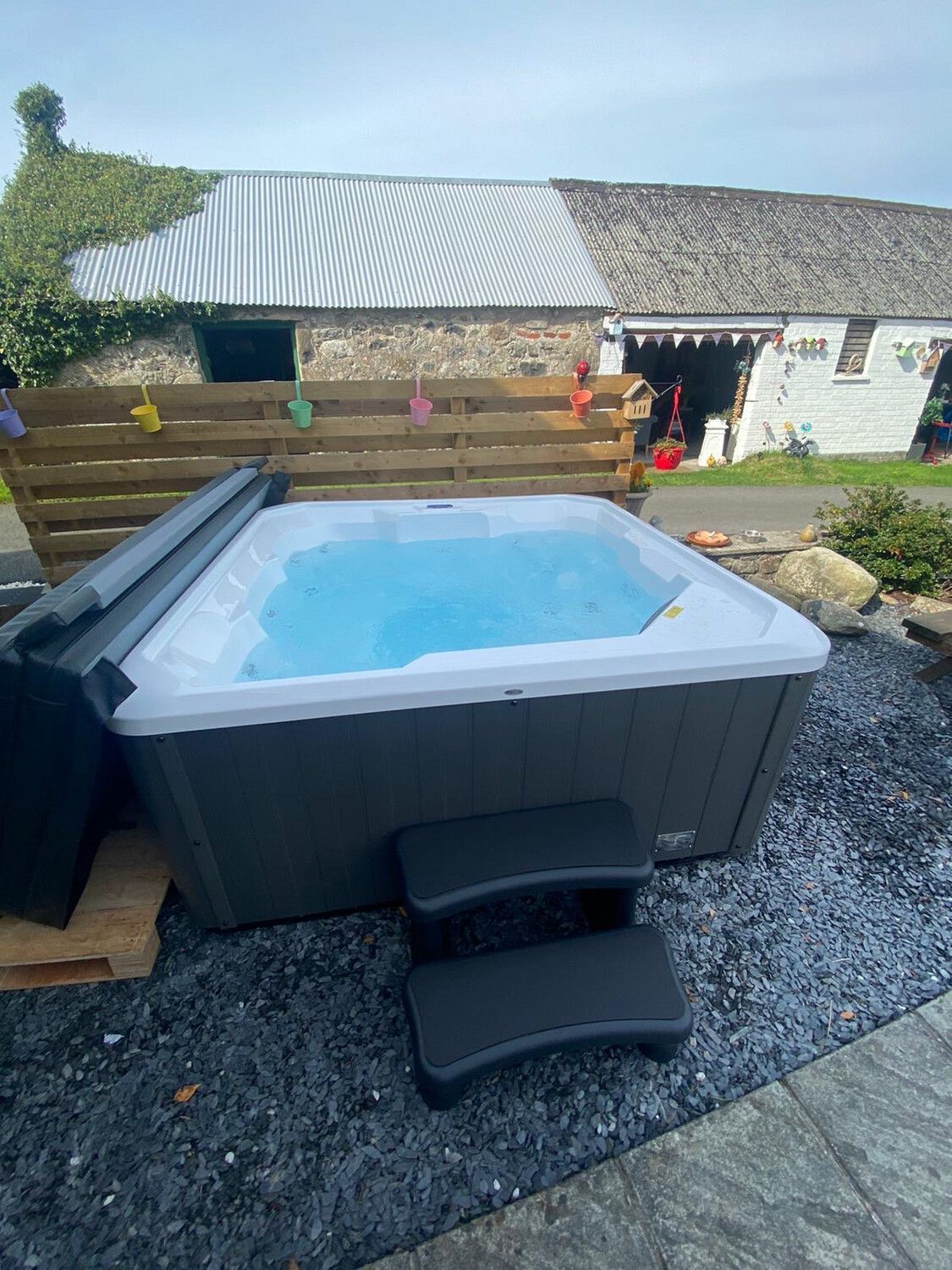 Hot tub with blue water, dark gray exterior, and black steps set on a gravel base. 