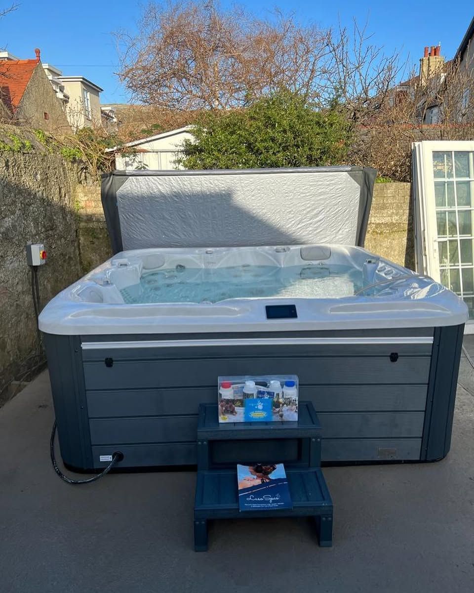 Hot tub with open lid, steps in front. Outdoor setting, gray siding.