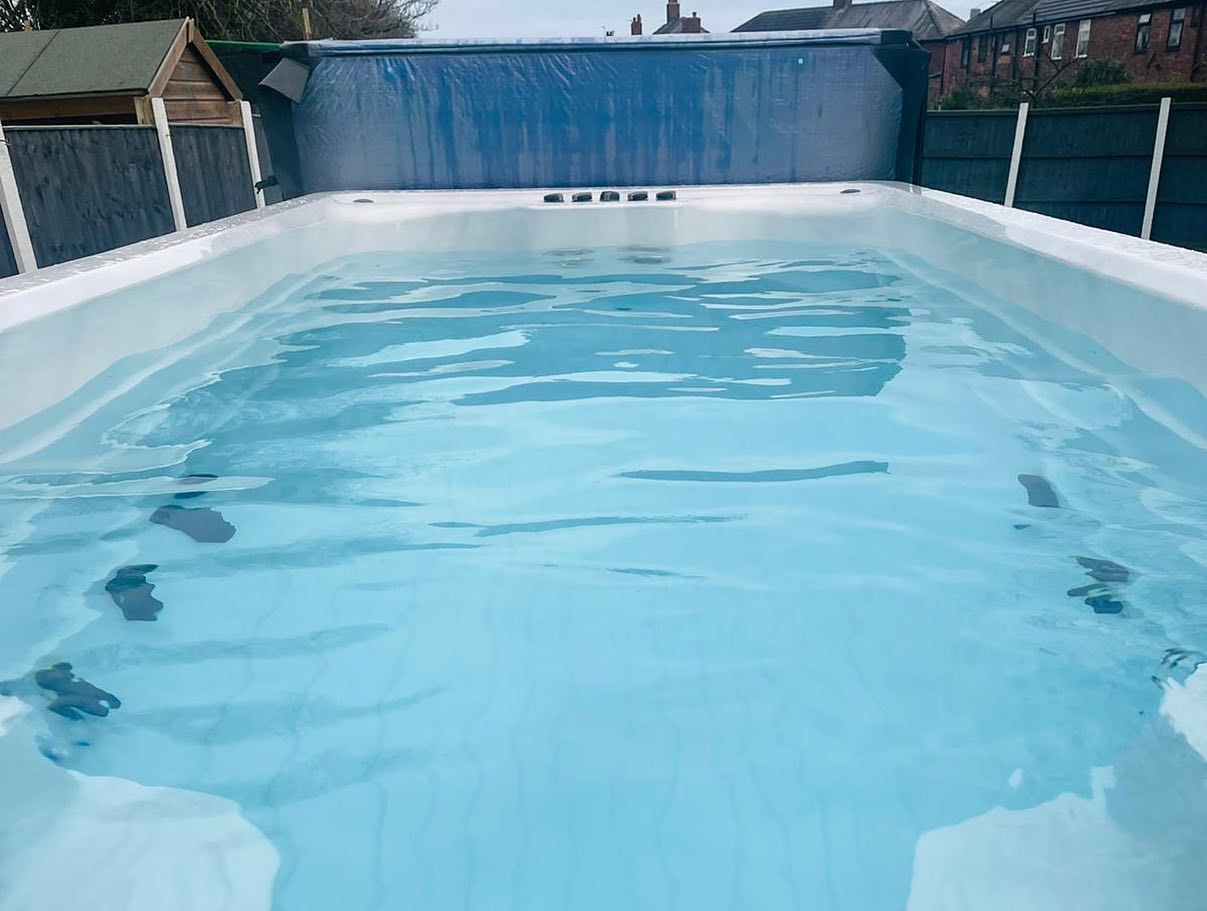 Rectangular, above-ground swimming pool filled with blue water. Blue tarp is at one end. Outdoors, overcast.