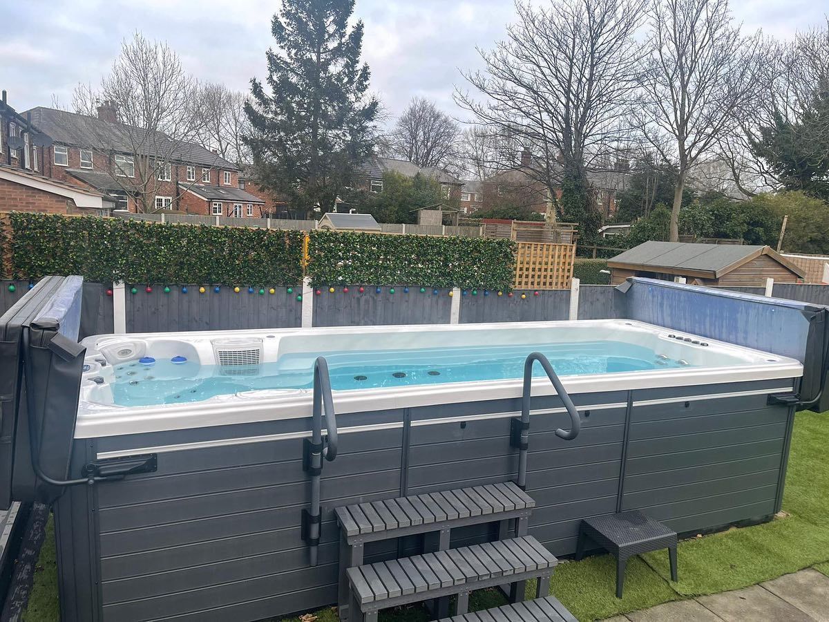A swim spa with gray exterior and steps in a backyard with a hedge and houses in the background.