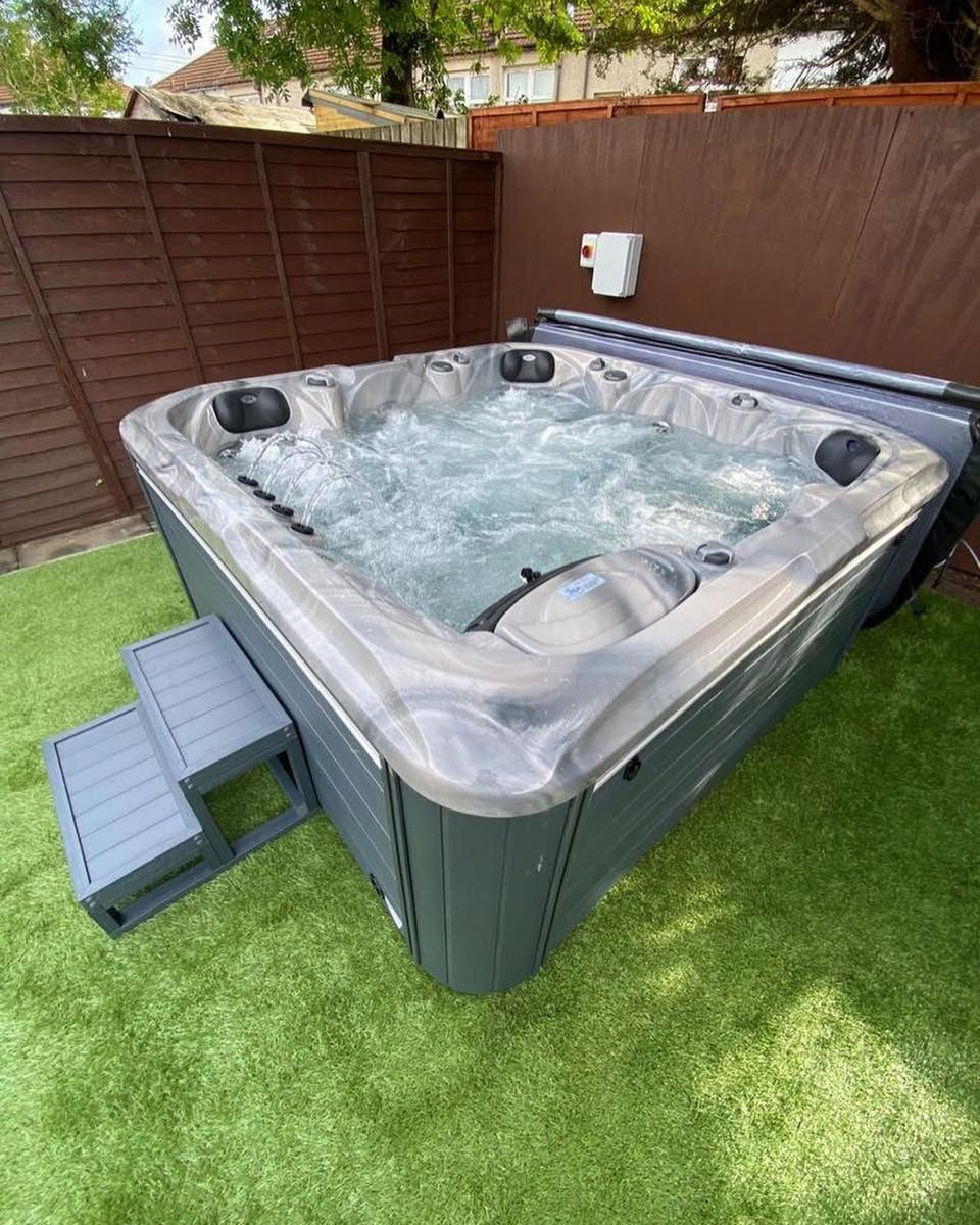 Hot tub with bubbling water on green turf, beside a brown fence, with gray steps.