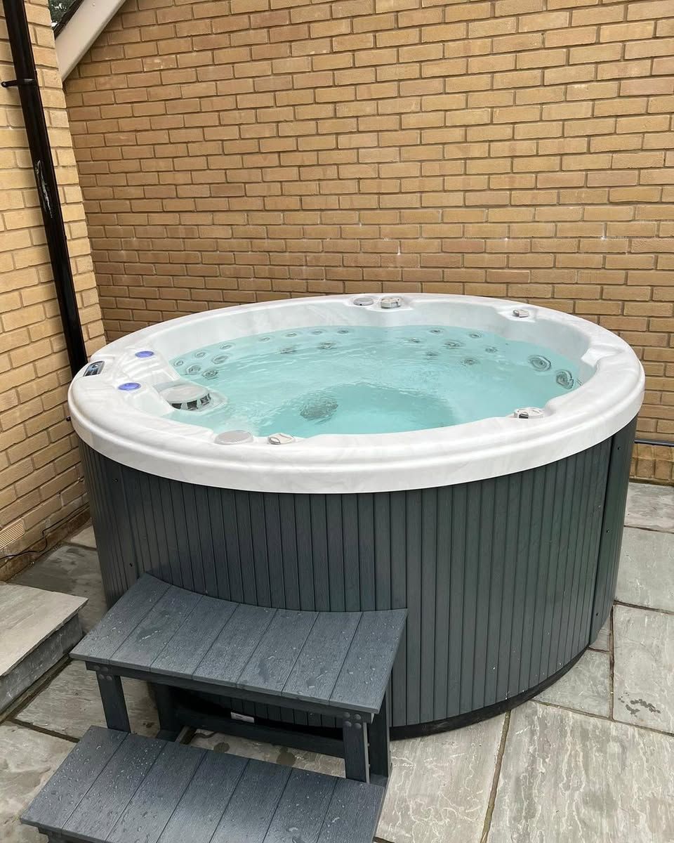 Round hot tub, grey exterior, filled with water, next to brick wall. Grey step stool in front.