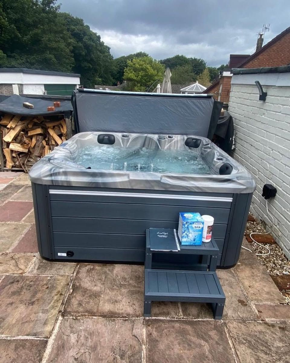 Hot tub on a patio with open cover, steps, and chemicals. Gray tub, overcast day.