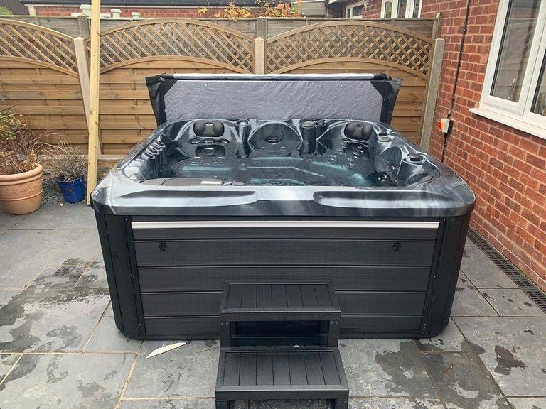 Black hot tub with open cover, two-step stool, on patio in front of a fence.