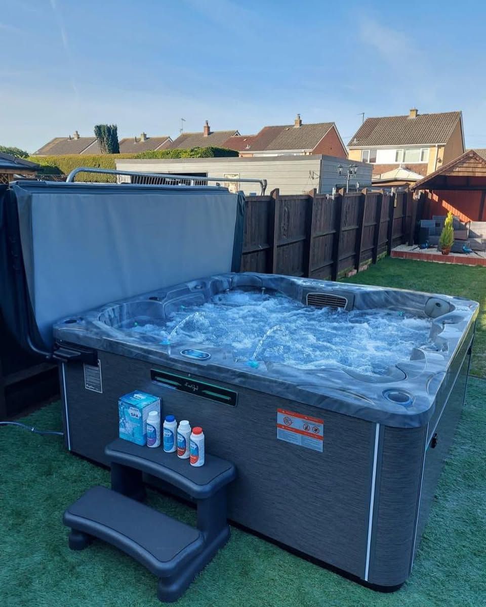 Hot tub in backyard with open cover, chemicals, and steps, bubbling water. Gray and blue tones.