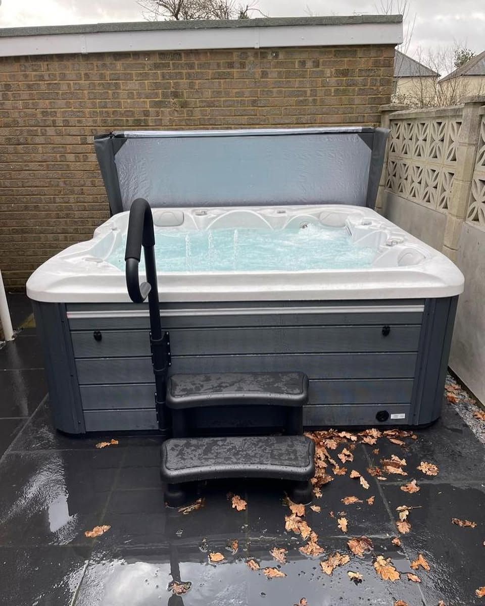 Hot tub with open cover, steps, and handrail outside against a brick wall. Gray and white colors.