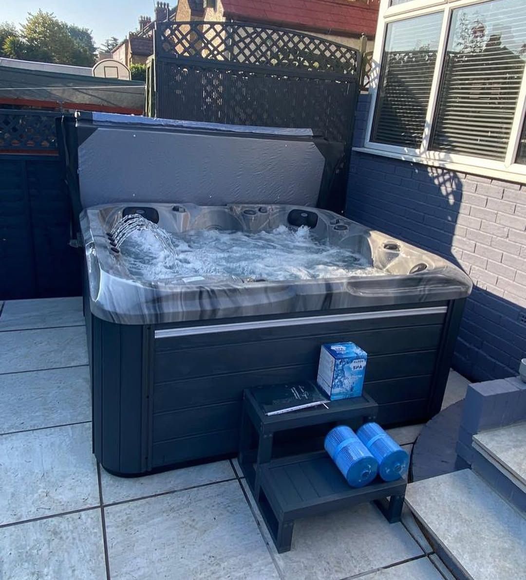 Hot tub on a patio with the cover open. Blue and gray tones, a small step, and chemicals are visible.