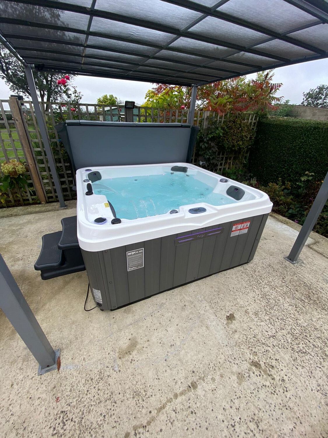 Hot tub under a canopy, on a concrete patio. Grey and white with water bubbling inside.