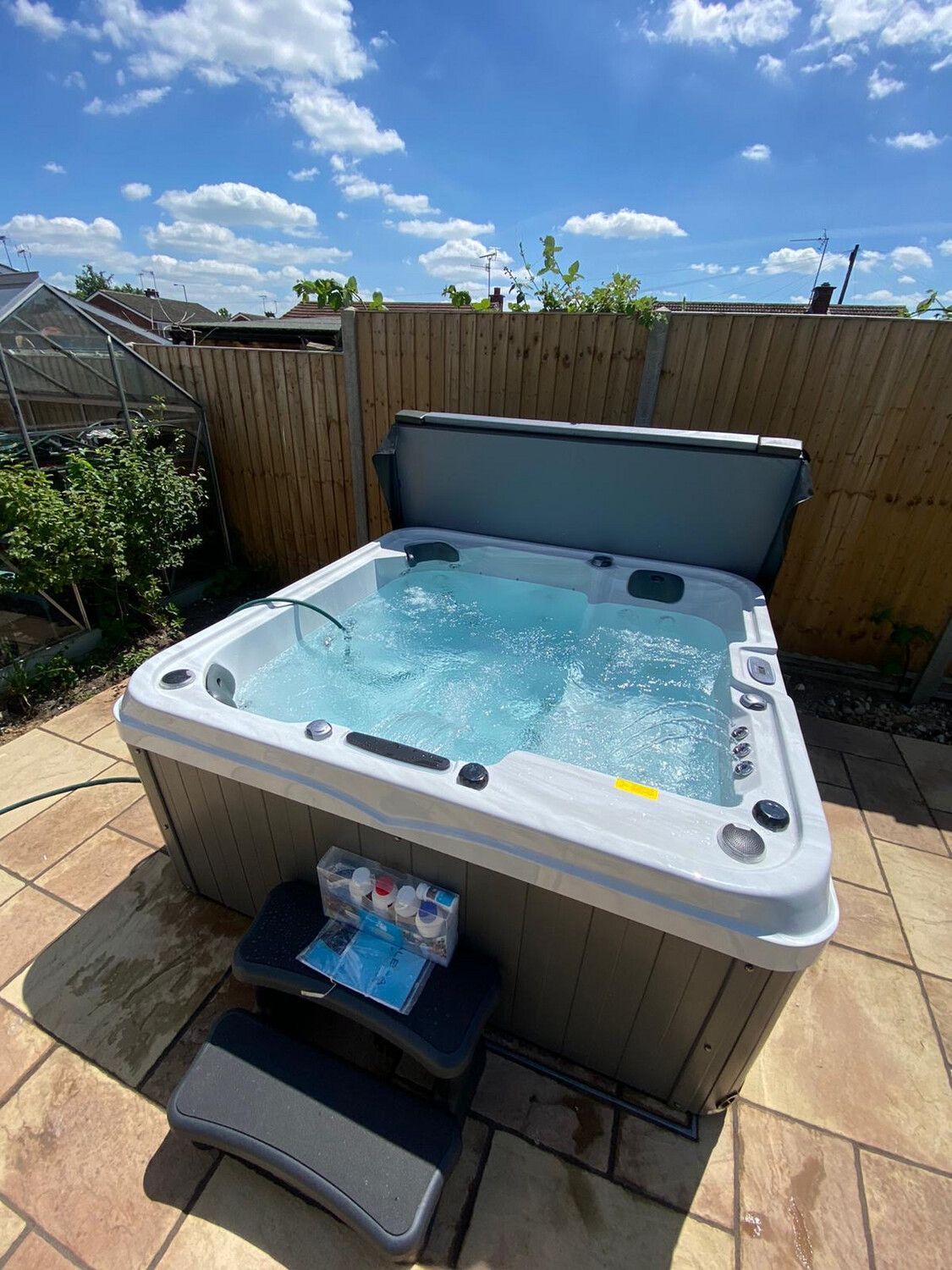 Hot tub on a stone patio with cover open; blue water and bubbles. Blue sky and fence in the background.