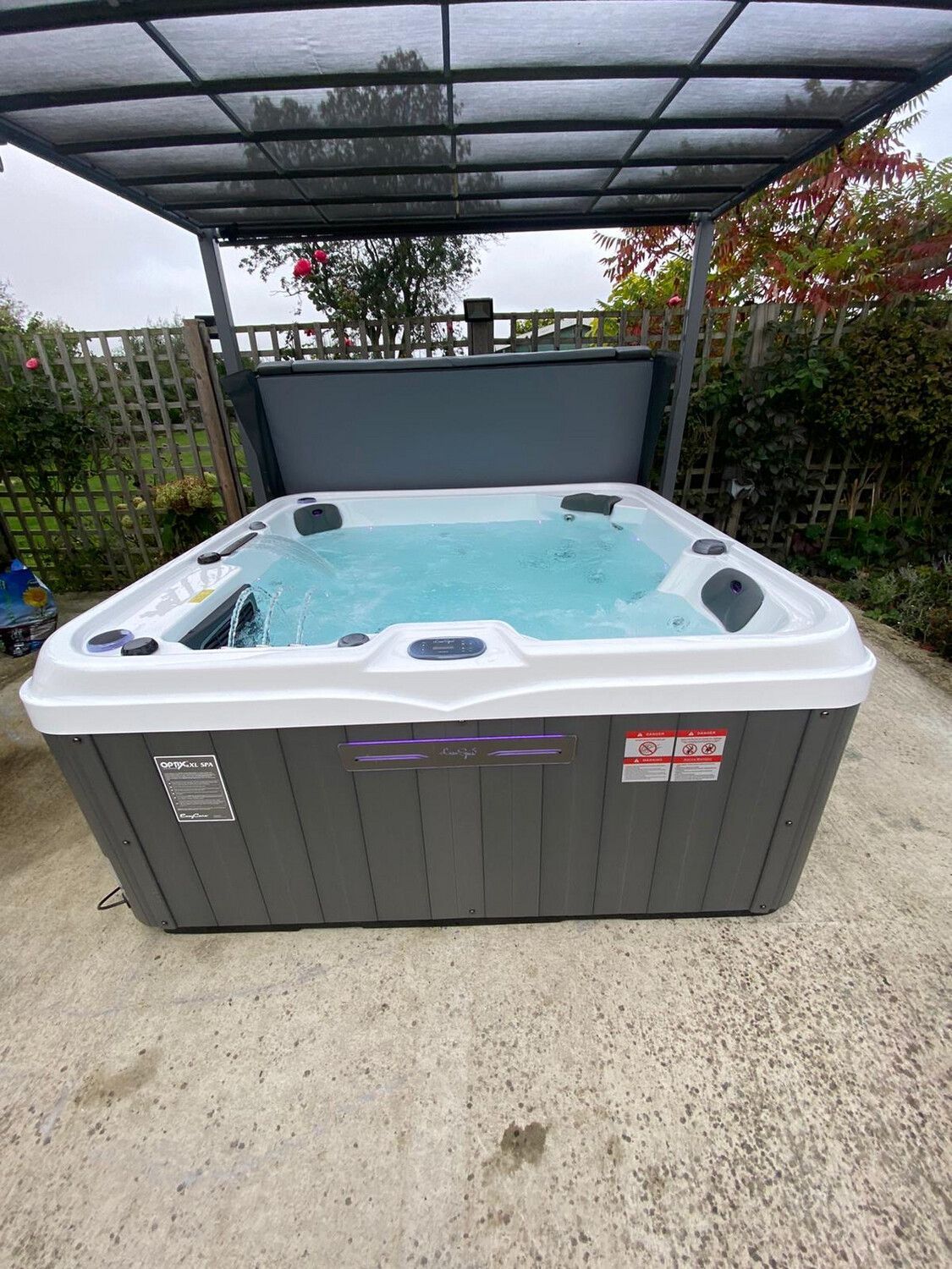 A square hot tub with open cover, filled with water under a canopy on a patio.