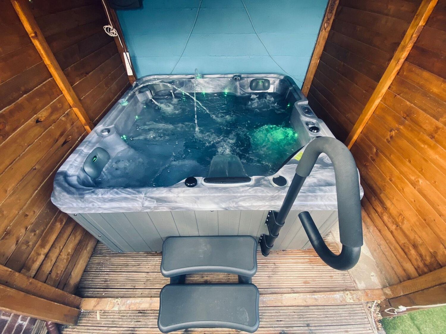 Hot tub in a wooden enclosure with steps and a handrail. Water is swirling with green lights.