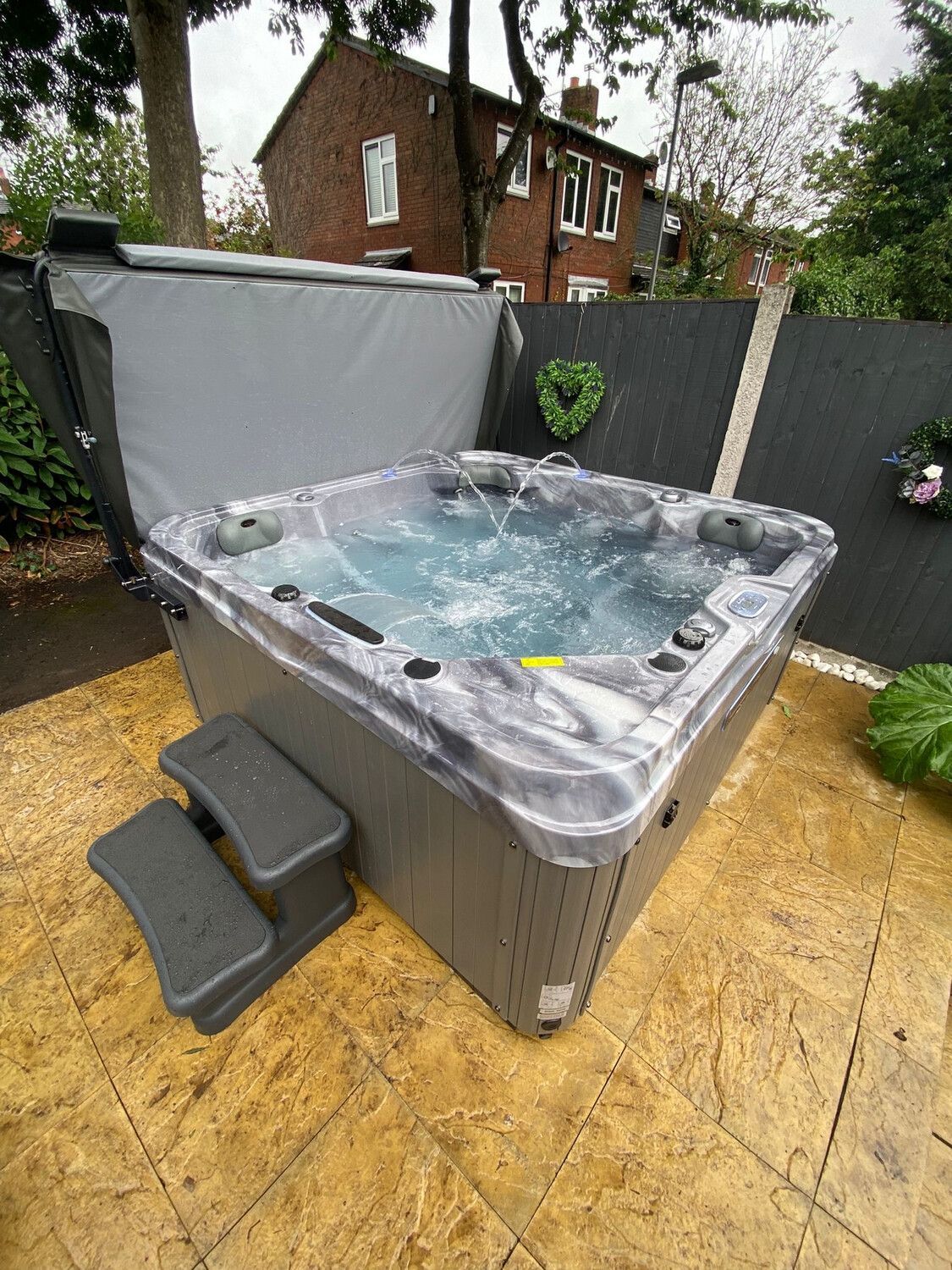 Hot tub with open cover, steps, and water jets outdoors on a patio with a fence and house in the background.