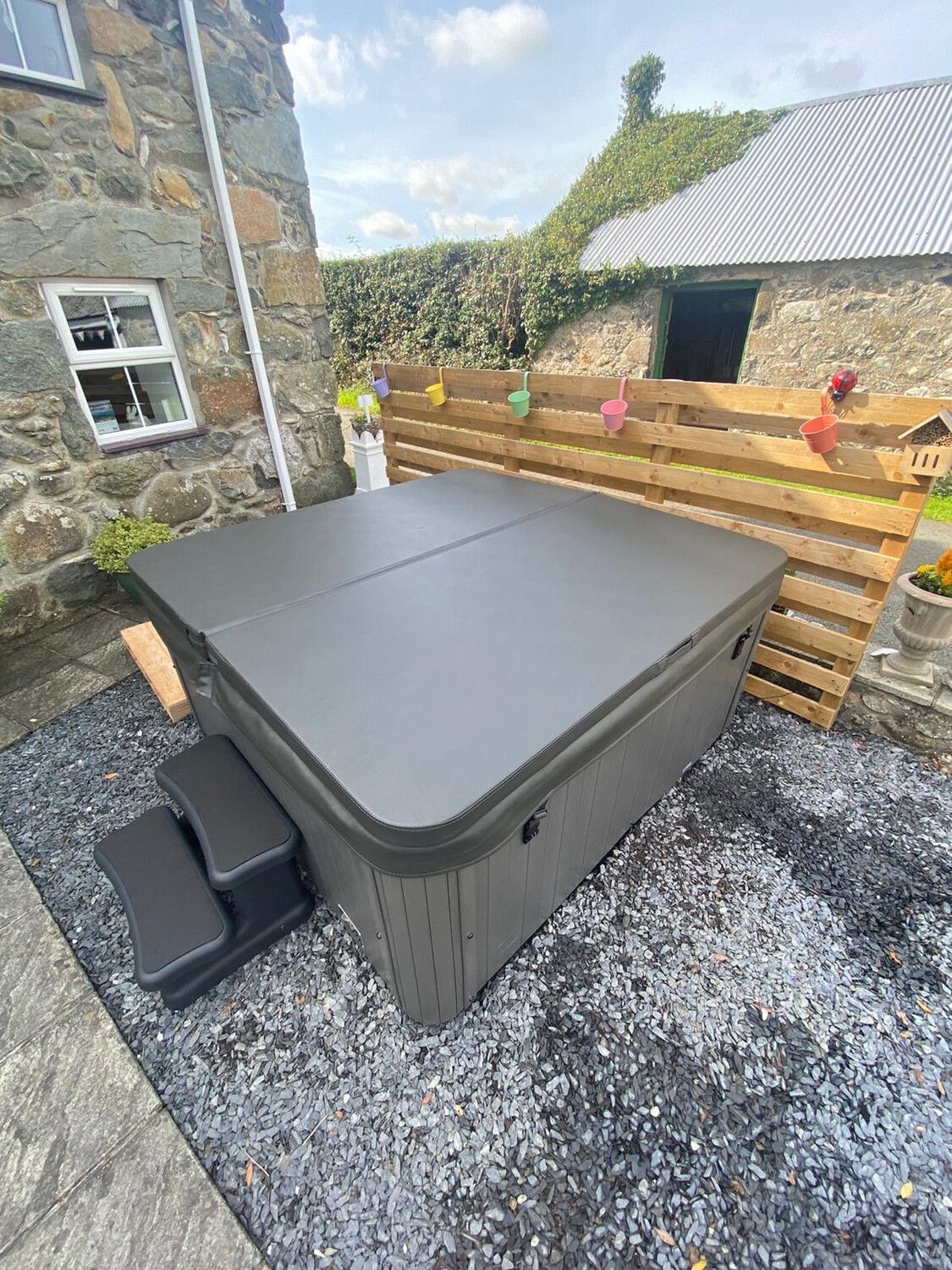 A gray hot tub with steps sits on gravel next to a stone building and wooden fence.