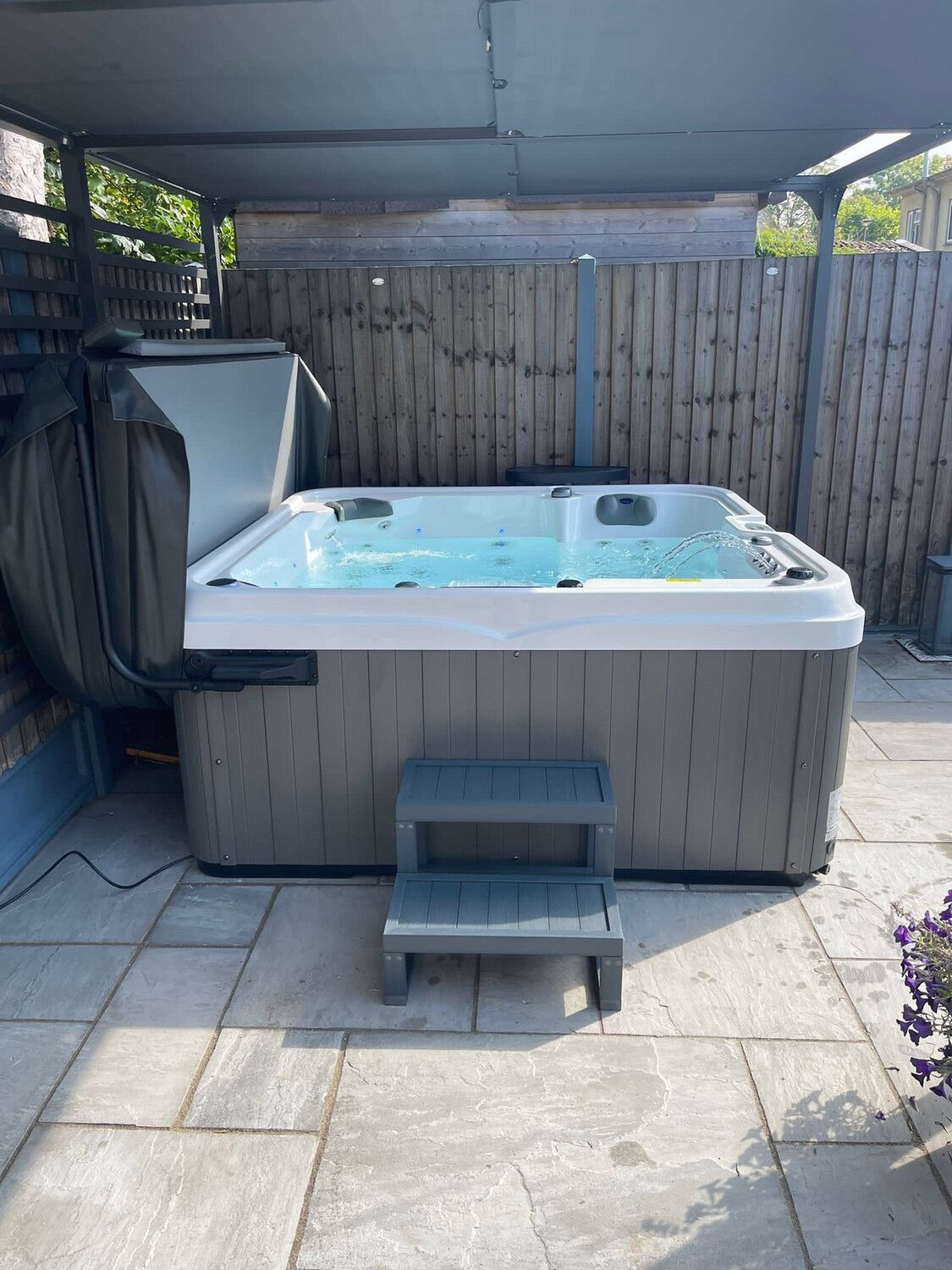 Hot tub with cover open, grey steps, and water, set outdoors under a canopy.