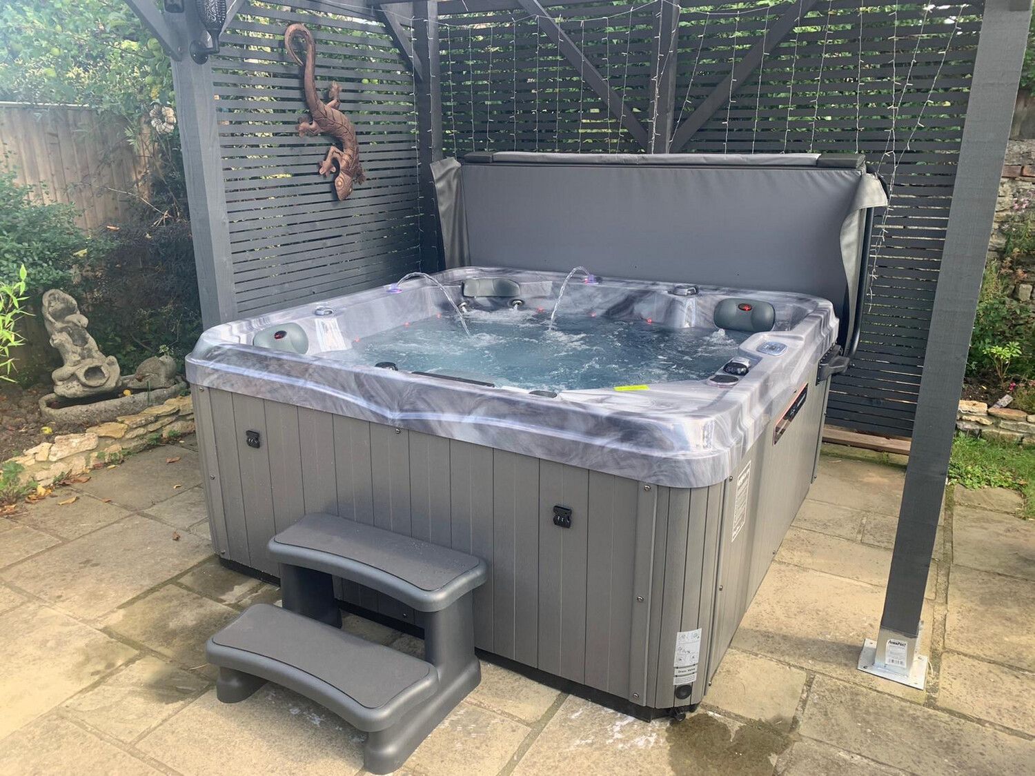 Gray hot tub with partially open cover, step stool, and gazebo in a backyard.