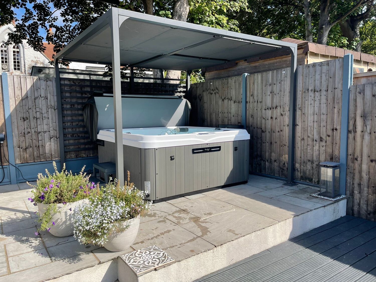 Hot tub under a gray canopy on a patio, surrounded by wooden fencing and potted plants.