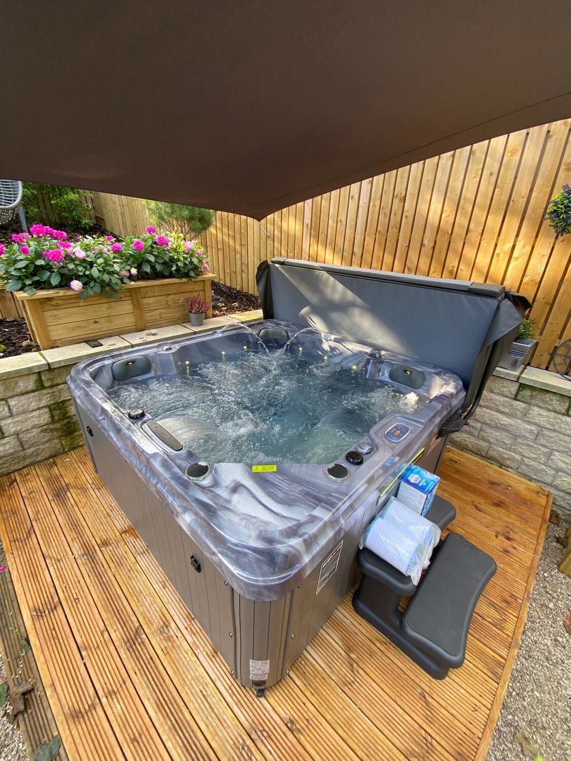 Hot tub on wooden deck, with cover open.  Brown shade canopy overhead.  Flowers, fence, and stone wall in the background.