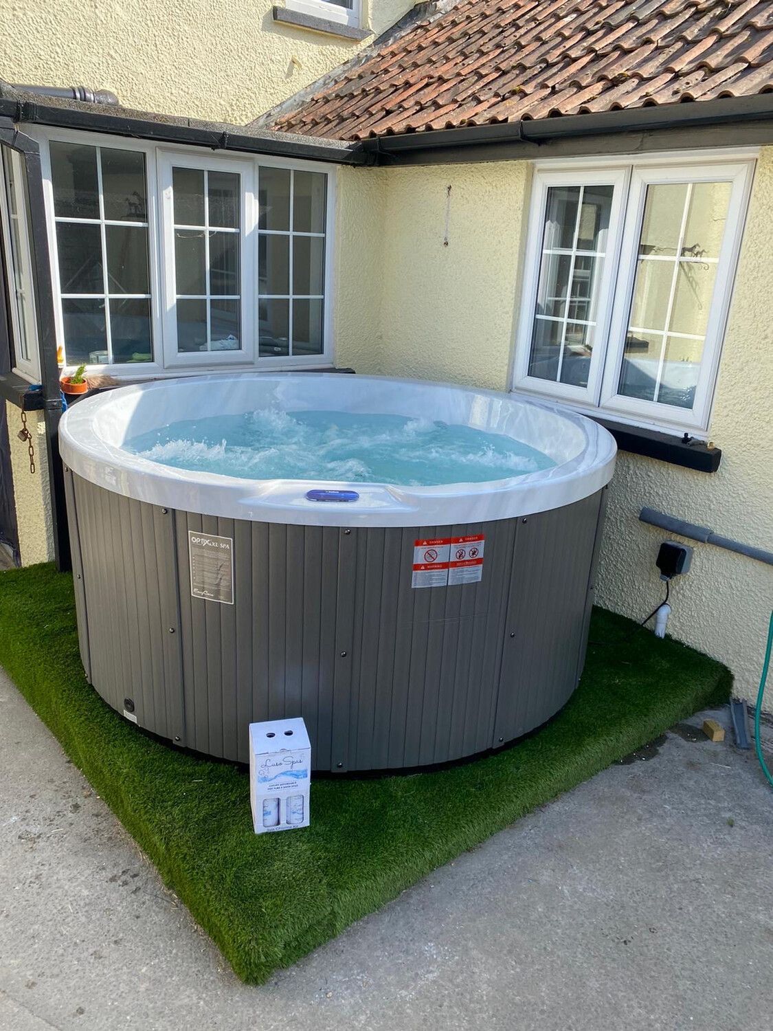 Hot tub on green artificial turf next to a building with windows and red-tiled roof.