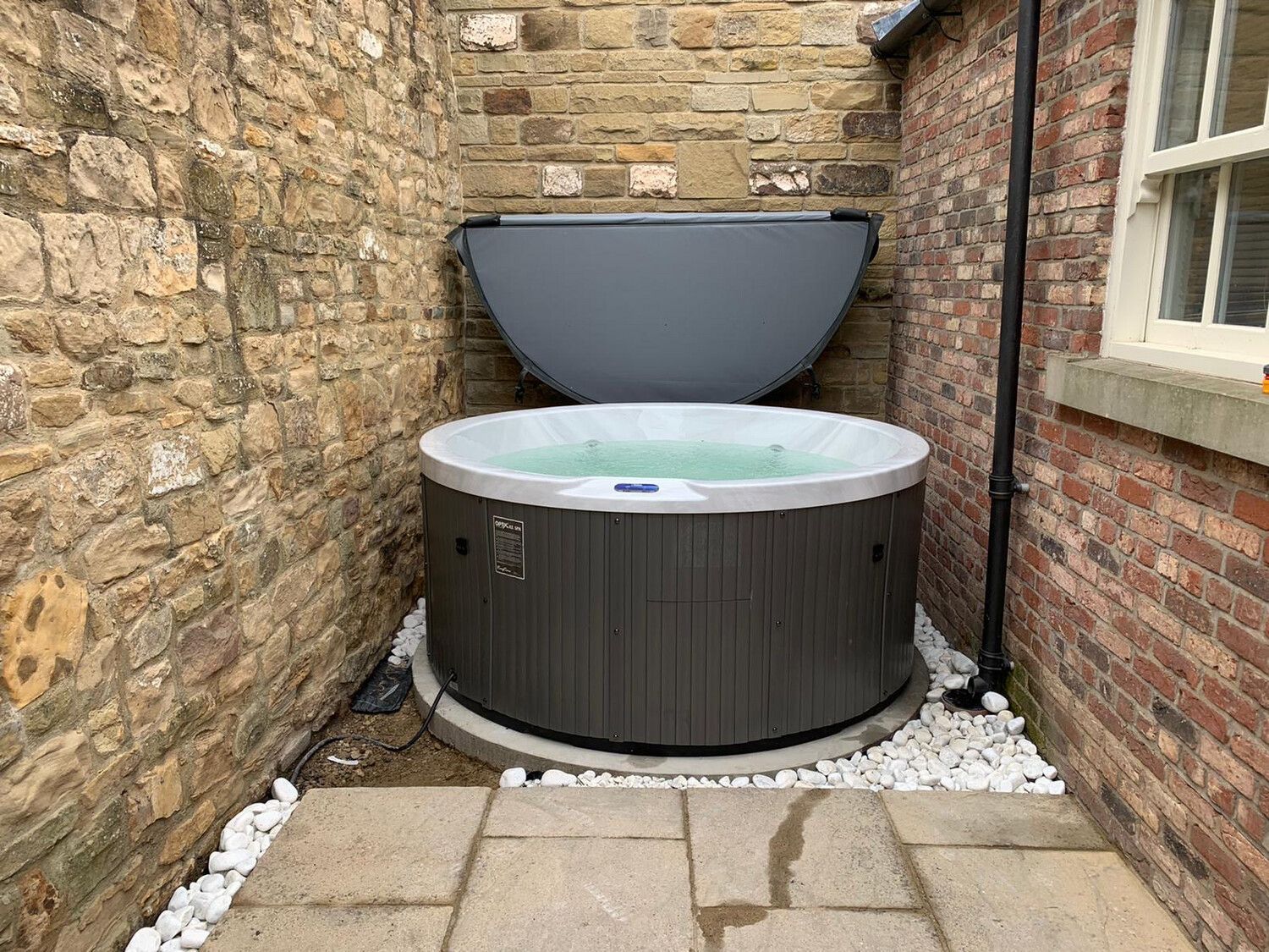 Hot tub in a small, stone-walled courtyard with its cover open, surrounded by white stones and paving.