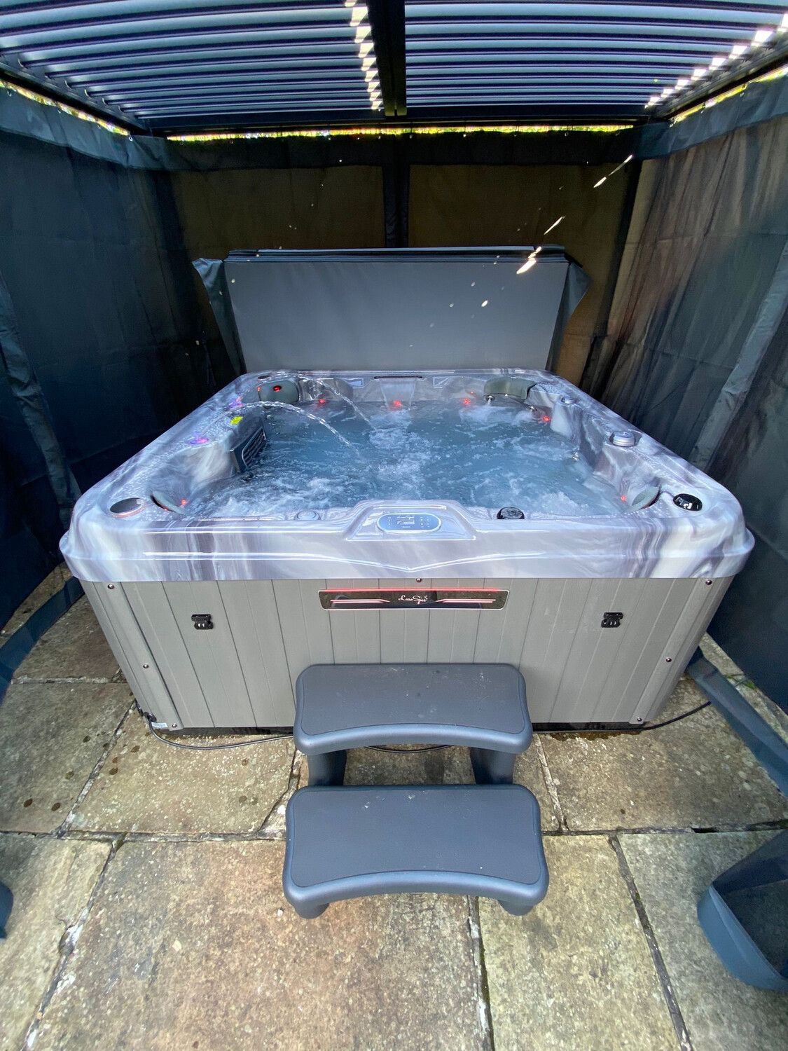 A bubbling hot tub with partially open cover, gray steps, set under a gazebo.